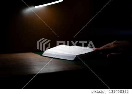 Person reading book at dimly lit desk, with light casting soft glow on pages. Evokes sense of quiet concentration, introspection, and peaceful atmosphere ideal for studying or reading 131458333