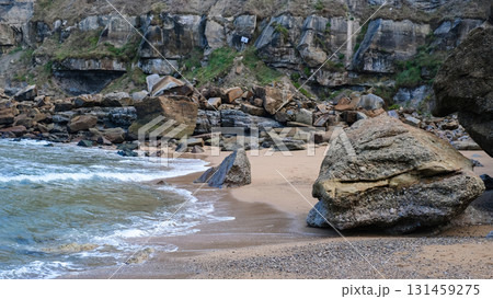 A wide, dramatic landscape photograph of a rugged seaside beach in Asturias, Spain, characterized by a steep, eroding cliff face covered in layered rock formations. A wide, dramatic landscape photograph of a rugged seaside beach in Asturias, Spain, characterized by a steep, eroding cliff face covered in layered rock formations. 131459275