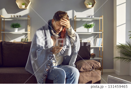 Man feeling cold sitting on sofa at home measuring temperature with thermometer Man feeling cold sitting on sofa at home measuring temperature with thermometer 131459465