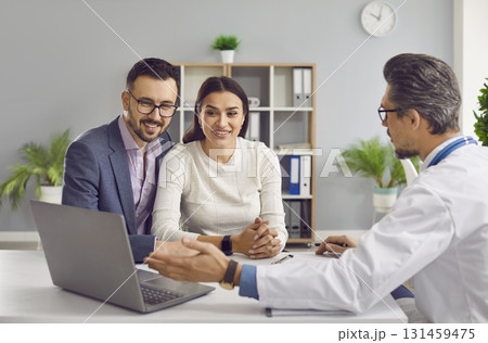 Doctor shows something on laptop while talking to happy couple planning children Doctor shows something on laptop while talking to happy couple planning children 131459475