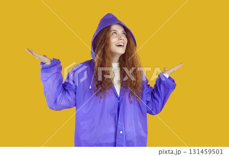 Happy young woman in purple raincoat looking up, spreading her hands and smiling Happy young woman in purple raincoat looking up, spreading her hands and smiling 131459501