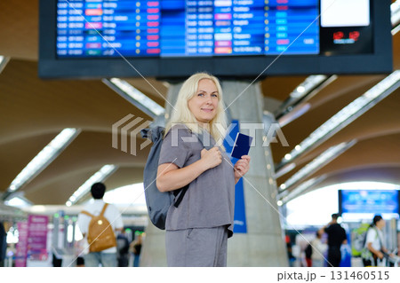 Woman with passport and backpack stands at airport terminal looking prepared for travel amid departure board and other travelers Woman with passport and backpack stands at airport terminal looking prepared for travel amid departure board and other travelers 131460515