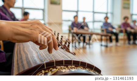 Close-up view of hands playing traditional Korean gayageum stringed instrument in bright music classroom with students sitting at tables in background during music lesson Close-up view of hands playing traditional Korean gayageum stringed instrument in bright music classroom with students sitting at tables in background during music lesson 131461193