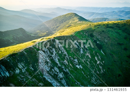 Aerial view of rugged mountain range with lush green slopes at sunset. Landscape showcases beauty of untouched nature with distant peaks and valleys bathed in soft sunlight. Carpathians, Chornogora. 131461935