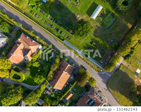 Aerial view of suburban homes with red roofs and lush greenery, separated from parallel garden plots and greenhouse by a paved road showcasing urban agriculture and residential harmony. Aerial view of suburban homes with red roofs and lush greenery, separated from parallel garden plots and greenhouse by a paved road showcasing urban agriculture and residential harmony. 131462143