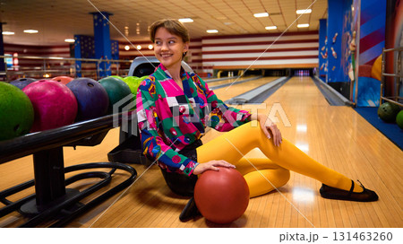 Woman in retro colorful outfit sitting by bowling balls on lane smiling 131463260