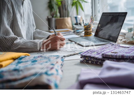 Young designer sketching clothes inspired by folk costumes with laptop and fabrics on table 131463747