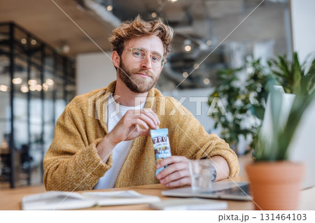 Man sitting at coworking desk eating plant based snack package labeled high protein zero sugar Man sitting at coworking desk eating plant based snack package labeled high protein zero sugar 131464103