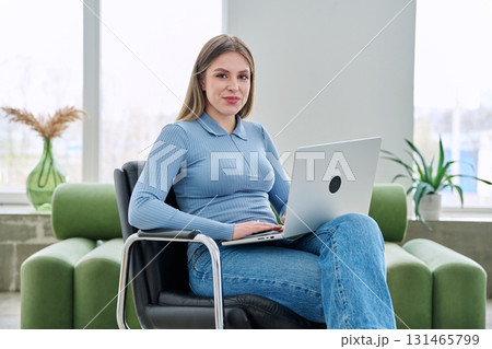 Portrait of young woman sitting on chair at home with laptop looking at camera Portrait of young woman sitting on chair at home with laptop looking at camera 131465799