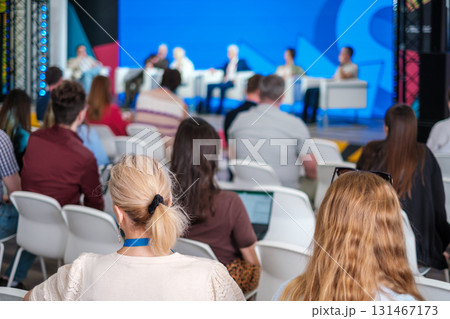 Audience watches a panel discussion on stage during a conference with a blue backdrop and bright lighting Audience watches a panel discussion on stage during a conference with a blue backdrop and bright lighting 131467173