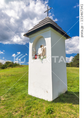 Religious shrine standing in a grassy field under a blue sky with clouds, topped with a cross, showcasing religious figurines and flowers inside. Spis region in Slovakia, Europe. 131467341