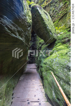 Adrspach-Teplice Rocks, sandstone formations in Hradec Kralove Region in the Czech Republic, Europe. A narrow wooden pathway through mossy rock formations, illuminated by light at the distant end. Adrspach-Teplice Rocks, sandstone formations in Hradec Kralove Region in the Czech Republic, Europe. A narrow wooden pathway through mossy rock formations, illuminated by light at the distant end. 131467404