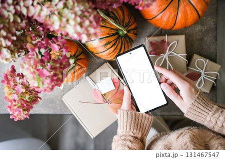 Top-down of person holding smartphone with blank white screen, pumpkins, fall leaves and gifts wrapped in craft paper. Perfect for app interfaces and e-commerce products during the fall holiday season 131467547