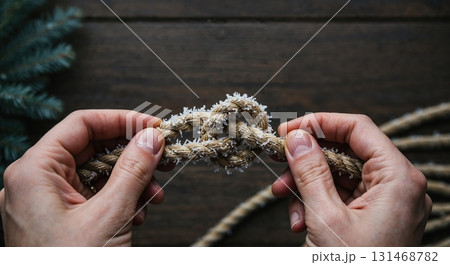 A close-up view of a person's hands carefully holding a thick rope with a central knot, all covered in delicate white frost against a dark wooden background and evergreen foliage. A close-up view of a person's hands carefully holding a thick rope with a central knot, all covered in delicate white frost against a dark wooden background and evergreen foliage. 131468782