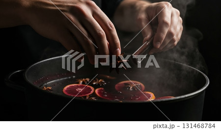 A person's hands carefully add star anise and a cinnamon stick to a steaming black pot filled with mulled wine and blood orange slices, set against a dark background with warm, dramatic lighting. A person's hands carefully add star anise and a cinnamon stick to a steaming black pot filled with mulled wine and blood orange slices, set against a dark background with warm, dramatic lighting. 131468784
