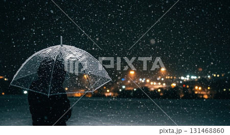 A person stands under a transparent umbrella during a heavy snowfall at night, with blurry city lights creating a warm bokeh background. 131468860