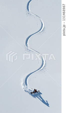 An aerial perspective showcases a person riding a specialized fat bike, carving an elegant S-shaped trail across a pristine white snowfield under clear winter sunlight. 131468987