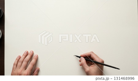 A top-down view shows a person's hands on a blank off-white paper, with one hand holding a black calligraphy pen ready to write or draw, alongside two small ink containers. A top-down view shows a person's hands on a blank off-white paper, with one hand holding a black calligraphy pen ready to write or draw, alongside two small ink containers. 131468996