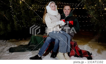 Winter evening warmth, young couple cuddling on blanket covered bench, holding steaming mugs, smiling amid gently falling snow and twinkling christmas lights 131471102