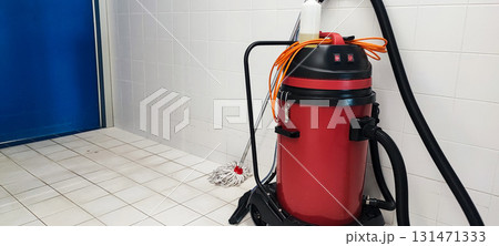 A red industrial vacuum cleaner with a mop and tools in a tiled gym restroom. Concept for hygiene, cleaning, and sanitation. A red industrial vacuum cleaner with a mop and tools in a tiled gym restroom. Concept for hygiene, cleaning, and sanitation. 131471333