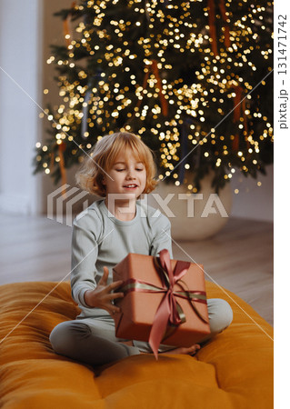 Smiling girl in pajamas tossing gift box near Christmas tree with lights in cozy room Smiling girl in pajamas tossing gift box near Christmas tree with lights in cozy room 131471742