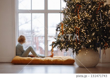 Girl sitting on cushion by window near decorated Christmas tree with lights in cozy room Girl sitting on cushion by window near decorated Christmas tree with lights in cozy room 131471743