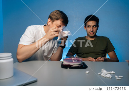 Two young men sitting at table with birthday cake, one snorting powdered drug while other watches, illustrating risks of youth substance abuse. Concept of party atmosphere with substance abuse. Two young men sitting at table with birthday cake, one snorting powdered drug while other watches, illustrating risks of youth substance abuse. Concept of party atmosphere with substance abuse. 131472458