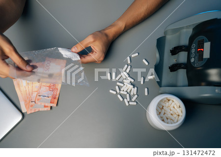 Drug dealer counting Russian rubles preparing drugs, pills, powder narcotics sitting at table with money counting machine in dark room, close-up, top view. Concept of drug dealing or addiction. Drug dealer counting Russian rubles preparing drugs, pills, powder narcotics sitting at table with money counting machine in dark room, close-up, top view. Concept of drug dealing or addiction. 131472472