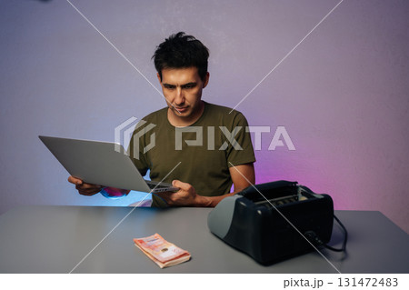 Portrait of young man sitting at desk, using laptop and money counter machine for detecting counterfeit banknotes, surrounded by vibrant purple background. Financial accounting in modern office. Portrait of young man sitting at desk, using laptop and money counter machine for detecting counterfeit banknotes, surrounded by vibrant purple background. Financial accounting in modern office. 131472483