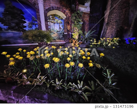 A serene night scene features a large, brightly lit artificial tree with colorful ornaments standing over a decorative, heart-shaped pond surrounded by illuminated foliage and pathways. 131473196