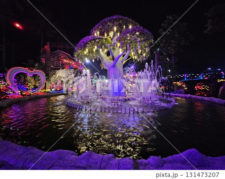 A stunning artificial tree adorned with countless colorful lights stands as the centerpiece of a vibrant fountain display, surrounded by more luminous decorations in a festive nighttime park. A stunning artificial tree adorned with countless colorful lights stands as the centerpiece of a vibrant fountain display, surrounded by more luminous decorations in a festive nighttime park. 131473207