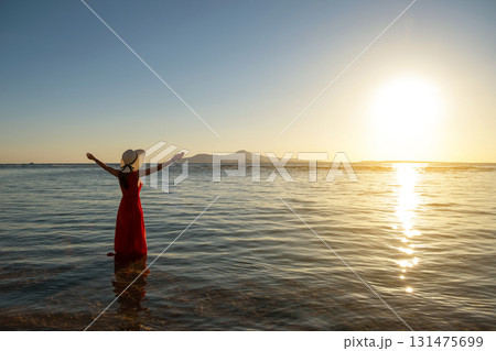 Young woman wearing long red dress and straw hat raising hands standing in sea water at the beach enjoying view of rising sun in early summer morning. Young woman wearing long red dress and straw hat raising hands standing in sea water at the beach enjoying view of rising sun in early summer morning. 131475699