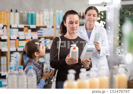 Woman with schoolgirl daughter are busy choosing cream at pharmacy. Pharmacist in background 131476797