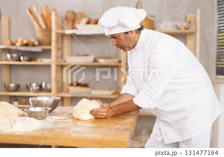 Male baker stands at work bench, kneading and shaping dough to make bread, croissants and baguettes 131477164