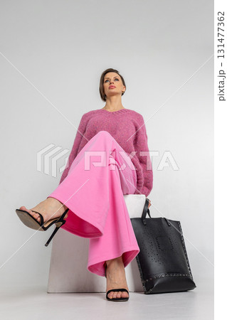 Portrait beautiful woman of model appearance. Girl in pink sweatshirt and trousers, black bag on grey background. Pretty female with trendy makeup posing in studio. Vertical photo. 131477362
