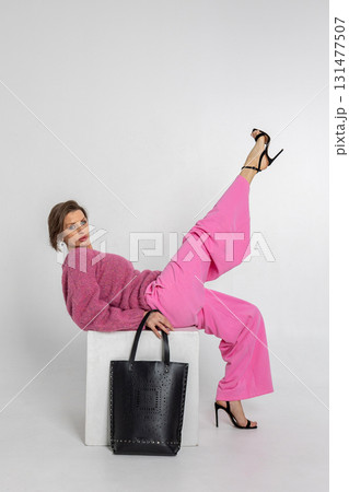 Portrait beautiful woman of model appearance. Girl in pink sweatshirt and trousers, black bag on grey background. Pretty female with trendy makeup posing in studio. Vertical photo. Portrait beautiful woman of model appearance. Girl in pink sweatshirt and trousers, black bag on grey background. Pretty female with trendy makeup posing in studio. Vertical photo. 131477507