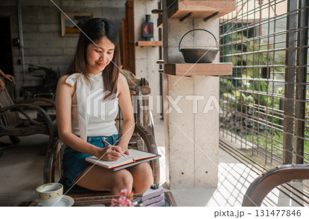 A young woman sits in a cozy cafe, writing in a notebook. The setting features rustic decor and ample natural light, creating a warm atmosphere. A young woman sits in a cozy cafe, writing in a notebook. The setting features rustic decor and ample natural light, creating a warm atmosphere. 131477846
