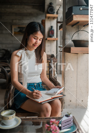 A young woman sits in a cozy cafe, writing in a notebook. The setting features rustic decor and natural light. A young woman sits in a cozy cafe, writing in a notebook. The setting features rustic decor and natural light. 131477847