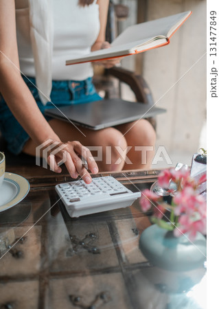 A woman calculates expenses while reading, surrounded by flowers and coffee, creating a cozy and productive atmosphere. 131477849