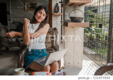 Young woman stretches while working on a laptop at home, surrounded by coffee and books, enjoying a cozy and productive atmosphere. Young woman stretches while working on a laptop at home, surrounded by coffee and books, enjoying a cozy and productive atmosphere. 131477850