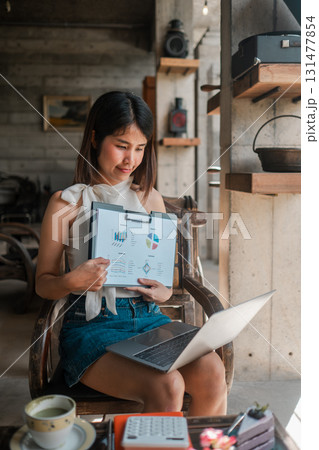 A young woman reviews business charts on a laptop in a cozy cafe, surrounded by coffee and a calculator. 131477854