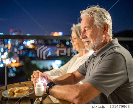 AI generated image of an elderly couple sitting on a balcony at night. The man smartwatch alerts him to take his medication. The peaceful city lights and modern setting create a calm atmosphere. 131479968