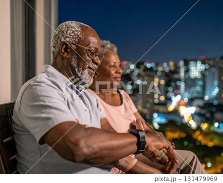 AI generated image of an elderly couple sitting on a balcony at night. The man smartwatch alerts him to take his medication. The peaceful city lights and modern setting create a calm atmosphere. 131479969