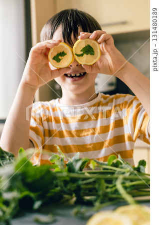 Smiling boy holding lemon slices with mint over eyes, funny playful moment in summer kitchen 131480189
