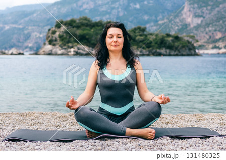 Young woman meditating in lotus pose on rocky beach with peaceful sea and mountain backdrop 131480325