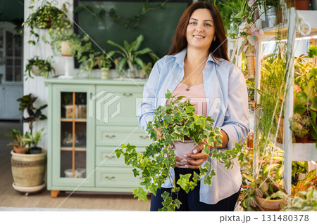Smiling woman holding hedera ivy potted plant at indoor garden retro home interior Smiling woman holding hedera ivy potted plant at indoor garden retro home interior 131480378