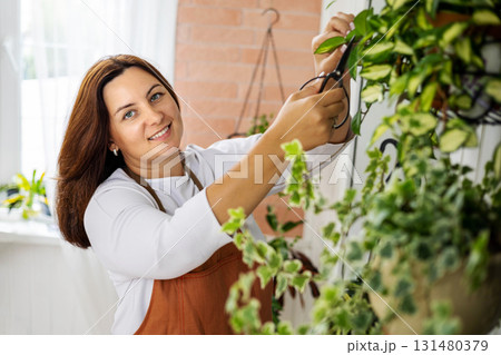 Portrait woman gardener in apron cutting branch of hedera ivy care cultivation at indoor home garden Portrait woman gardener in apron cutting branch of hedera ivy care cultivation at indoor home garden 131480379