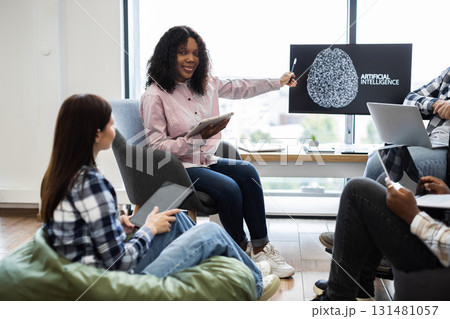 Young African woman presenting artificial intelligence project to colleagues. Group collaborates on project using technology such as laptops and tablets in cozy modern office. Young African woman presenting artificial intelligence project to colleagues. Group collaborates on project using technology such as laptops and tablets in cozy modern office. 131481057