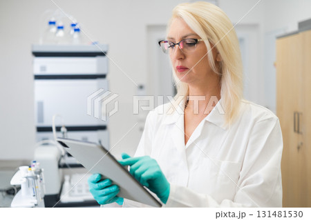 Middle aged female chemist using a tablet in a biochemistry laboratory. HPLC instrument in the background symbolizes accuracy and modern methods. 131481530