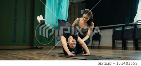 Female trainer helping student with upside-down yoga fly position in studio Female trainer helping student with upside-down yoga fly position in studio 131481555
